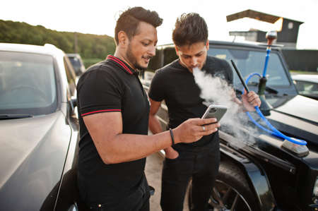 Two asian brothers man wear on all black posed near suv cars and smoke hookah.