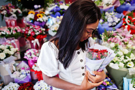 Beautiful african american girl in tender white dress with bouquet flowers in hands standing against...