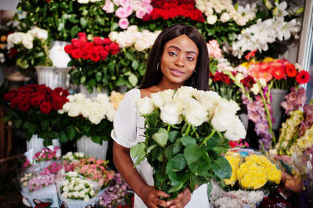 Beautiful african american girl in tender white dress with bouquet flowers in hands standing against floral background in flower shop.Black female florist.の写真素材