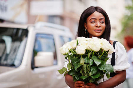 Beautiful african american girl holding bouquet of white roses flowers on dating in the city. Black businesswoman with bunch of flowers near car.の写真素材