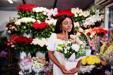 Beautiful african american girl in tender white dress with bouquet flowers in hands standing against floral background in flower shop.Black female florist.の写真素材