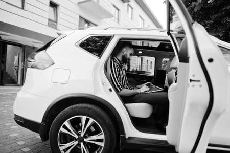 Successful arab man wear in striped shirt and sunglasses pose inside white suv car with laptop in hands.の写真素材