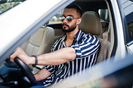 Successful arab man wear in striped shirt and sunglasses pose behind the wheel of his white suv car. Stylish arabian men in transport.の写真素材