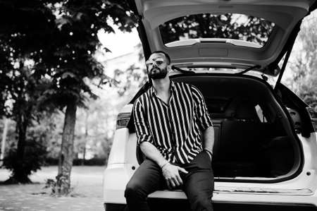 Successful arab man wear in striped shirt and sunglasses sitting in the trunk of his white suv car....