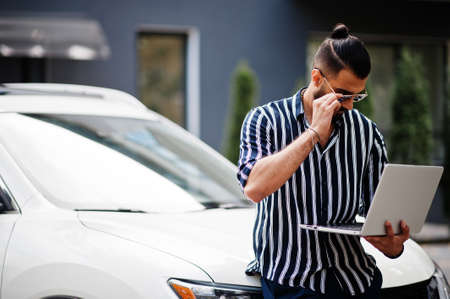 Successful arab man wear in striped shirt and sunglasses sitting on the roof of his white suv car.の写真素材