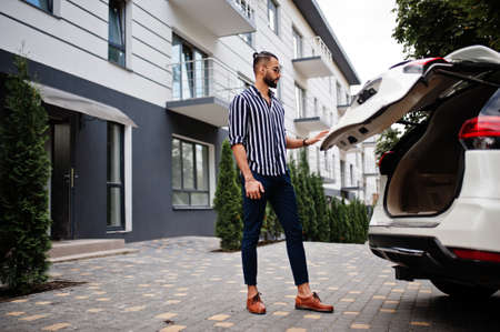 Successful arab man wear in striped shirt and sunglasses sitting on the roof of his white suv car.の写真素材