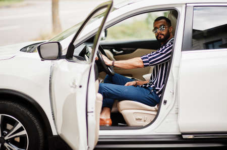 Successful arab man wear in striped shirt and sunglasses sitting on the roof of his white suv car.の写真素材