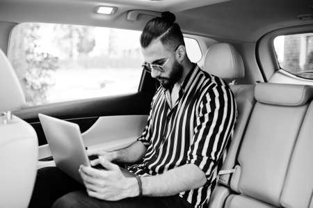 Successful arab man wear in striped shirt and sunglasses sitting on the roof of his white suv car.の写真素材