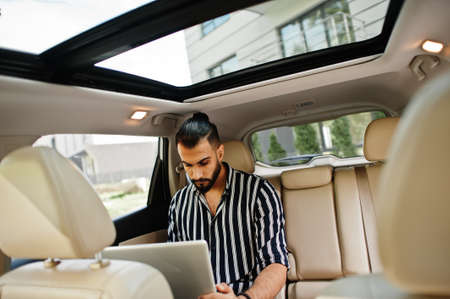 Successful arab man wear in striped shirt and sunglasses sitting on the roof of his white suv car.