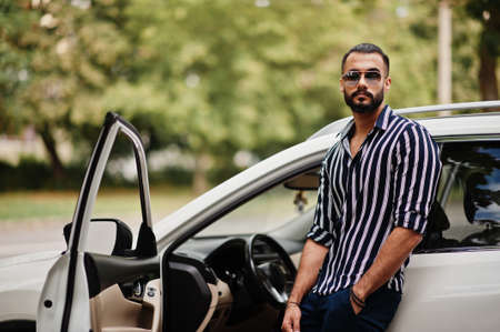 Successful arab man wear in striped shirt and sunglasses sitting on the roof of his white suv car.の写真素材