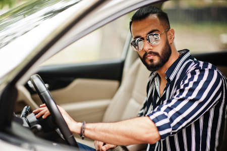 Successful arab man wear in striped shirt and sunglasses sitting on the roof of his white suv car.