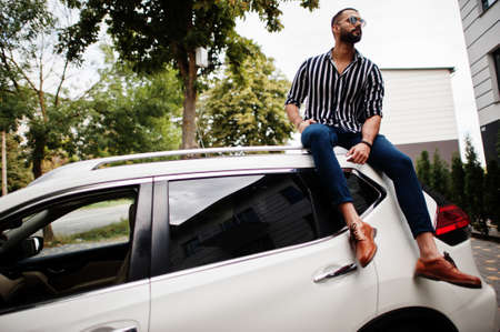 Successful arab man wear in striped shirt and sunglasses sitting on the roof of his white suv car.の写真素材
