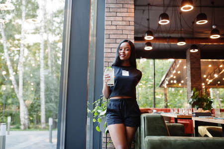 Fashionable feminist african american woman wear in black t-shirt and shorts, posed at restaurant with lemonade glass.の写真素材