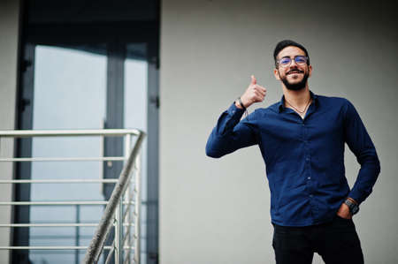 Middle eastern entrepreneur wear blue shirt, eyeglasses against office building and show thumb up.の写真素材