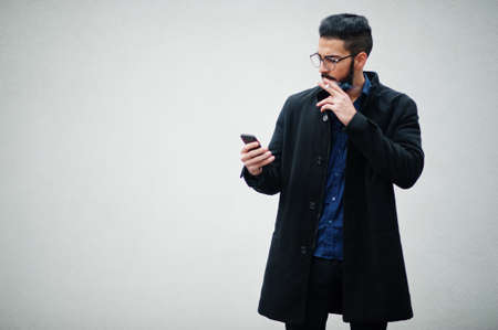 Middle eastern entrepreneur wear black coat and blue shirt, eyeglasses standing against white wall,...