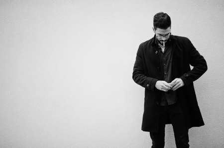Middle eastern entrepreneur wear black coat and blue shirt, eyeglasses standing against white wall and smoking cigarette.の写真素材