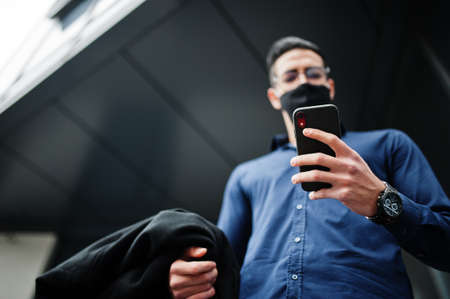 Middle eastern entrepreneur wear black face mask and eyeglasses look at mobile phone. New normal life after coronavirus pandemia.の写真素材
