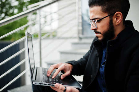 Middle eastern entrepreneur wear black coat and blue shirt, eyeglasses against office building with laptop.の写真素材