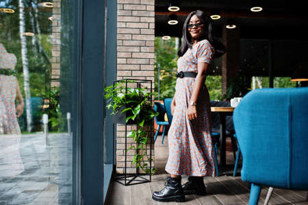 Fashion portrait of african american woman in long dress, boots and sunglasses indoor.