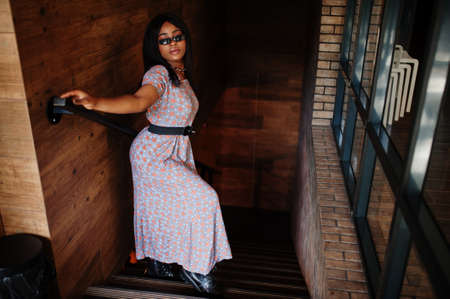 Fashion portrait of african american woman in long dress, boots and sunglasses indoor.