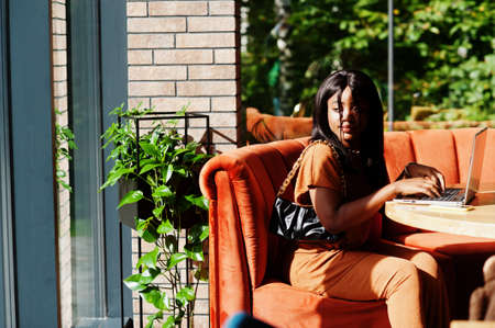Portrait of beauty young black woman, wear orange outfit, sitting with laptop on office.の写真素材