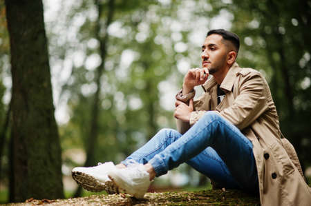 Stylish kuwaiti man at trench coat sit on tree at wood.の写真素材