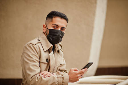 Stylish kuwaiti man at trench coat wear black face mask during covid pandemic with telephone.