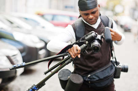 Young professional african american videographer holding professional camera with tripod pro equipment. Afro cameraman wearing black duraq making a videos.の写真素材