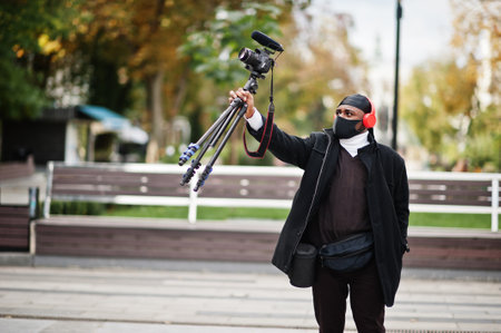 Young professional african american videographer holding professional camera with pro equipment. Afro cameraman wearing black duraq and face protect mask, making a videos.の写真素材