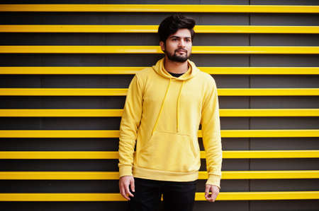 Urban young hipster indian man in a fashionable yellow sweatshirt. Cool south asian guy wear hoodie against striped background.の写真素材