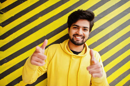 Urban young hipster indian man in a fashionable yellow sweatshirt. Cool south asian guy wear hoodie against striped background show fingers to camera.の写真素材