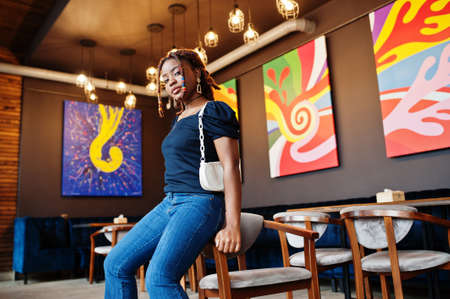 Lovely african american woman with dreadlocks in blue stylish jeans at cafe. Beautiful cool fashionable black young girl indoor.の写真素材