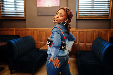Lovely african american woman with dreadlocks in blue stylish jeans jacket at cafe. Beautiful cool...