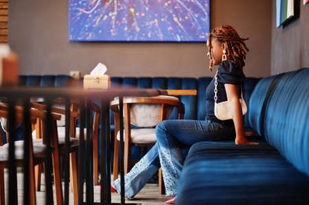 Lovely african american woman with dreadlocks in blue stylish jeans at cafe. Beautiful cool fashionable black young girl indoor.の写真素材