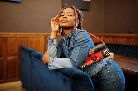 Lovely african american woman with dreadlocks in blue stylish jeans jacket at cafe. Beautiful cool...