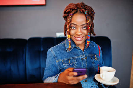 Lovely african american woman with dreadlocks in blue stylish jeans jacket at cafe hold mobile...