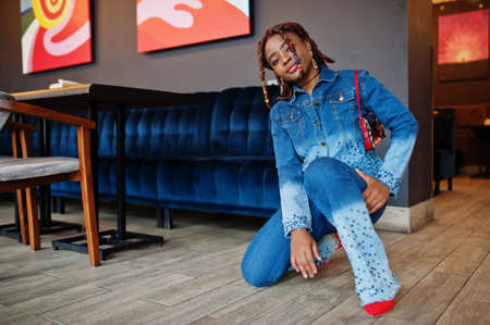 Lovely african american woman with dreadlocks in blue stylish jeans jacket at cafe. Beautiful cool...