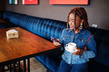 Lovely african american woman with dreadlocks in blue stylish jeans jacket at cafe hold mobile...