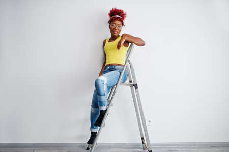 African american woman in yellow singlet and eyglasses against white wall stand om iron ladder.の写真素材