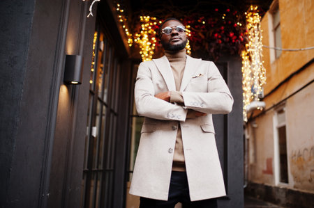 Stylish african american man wear beige jacket with sunglasses pose against house with golden...