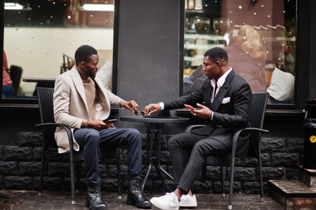 Two fashion black men sitting outdoor with mobile phone. Fashionable portrait of african american male models. Wear suit, coat and hat.の写真素材