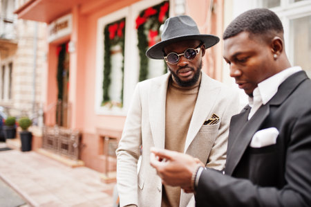 Two fashion black men stand near business car and look at cell phone. Fashionable portrait of african american male models. Wear suit, coat and hat.の写真素材