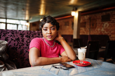 Urban young african american woman in pink top sit in cafe and drink latte coffee.
