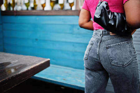Urban young african american woman in pink top and gray jeans with handbag. Afro fashion chic women back buttocks.の写真素材