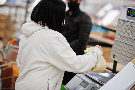 Asian couple wear in protective face mask shopping together in supermarket during pandemic. Weigh the goods.の写真素材