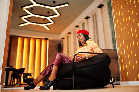 Elegant afro american woman in red french beret, big gold neck chain polka dot blouse and leather pants sit on pouf.の写真素材