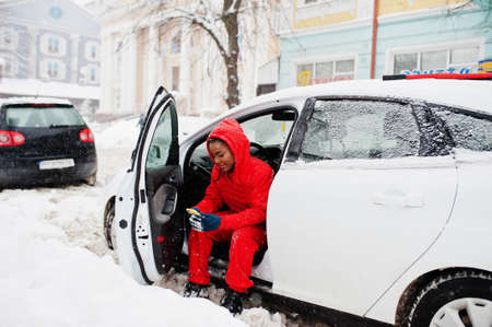 African american woman in red hoodie sitting inside car at winter snowy day with mobile phone.の写真素材