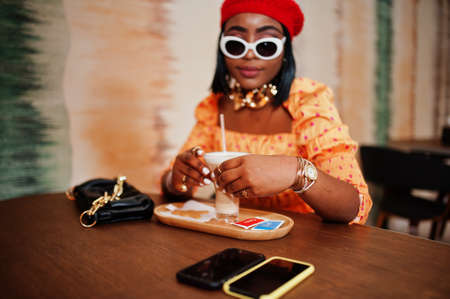 Elegant afro american woman in red french beret, big gold neck chain polka dot blouse pose indoor, sitting at restaurant and drink latte.の写真素材