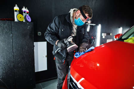 Car detailing concept. Man in face mask with orbital polisher in repair shop polishing orange suv car.の写真素材