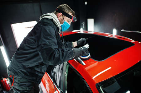 Car detailing concept. Man in face mask with orbital polisher in repair shop polishing roof of orange suv car.の写真素材